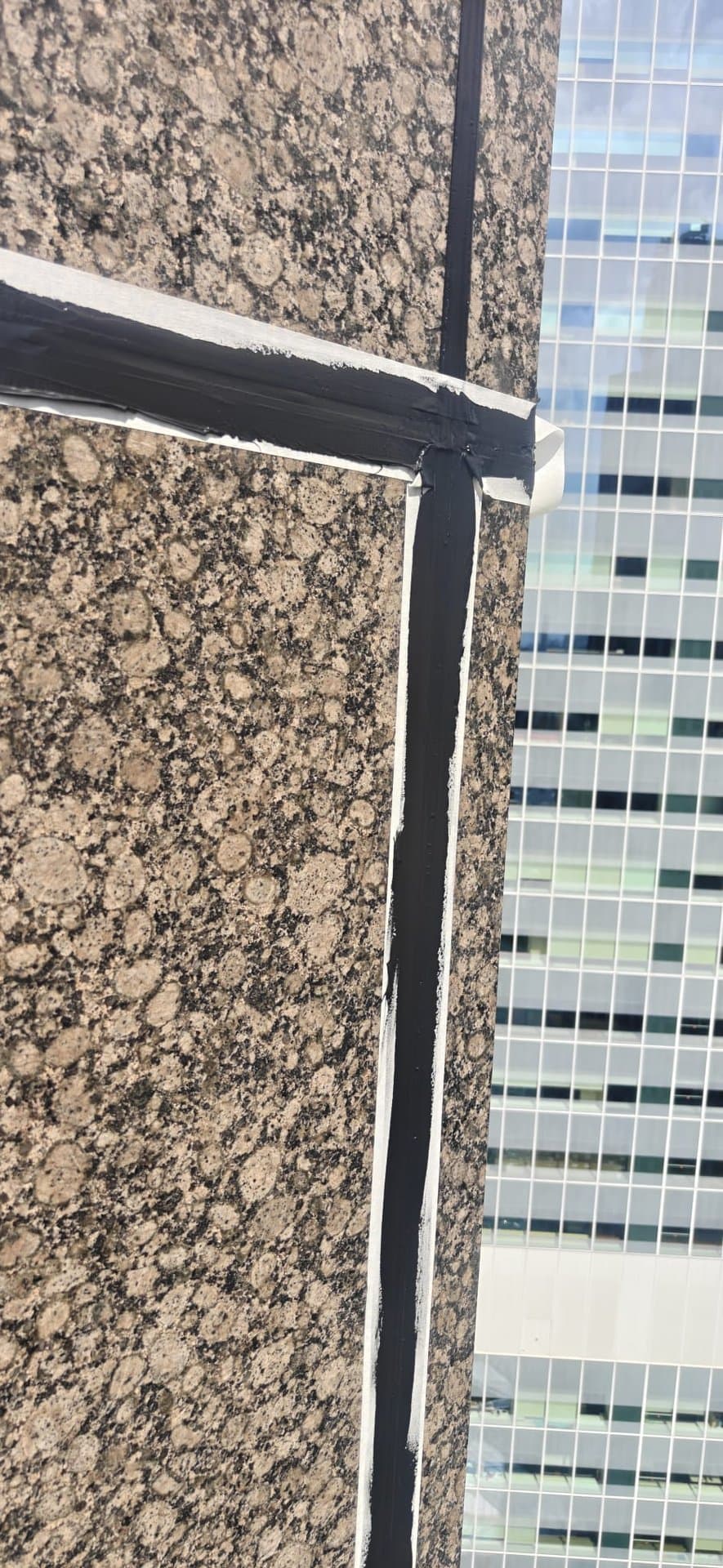 Hydroseal façade sealant joint with fresh masking tape applied to granite cladding during rope-access waterproofing works on an office tower in Singapore’s CBD