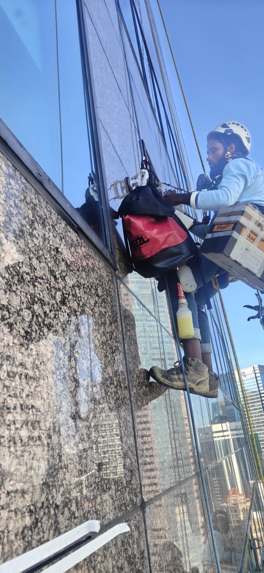 Hydroseal IRATA rope-access technician mid-abseil working a granite-and-glass façade joint during sealant replacement on a high-rise office building in Singapore’s CBD