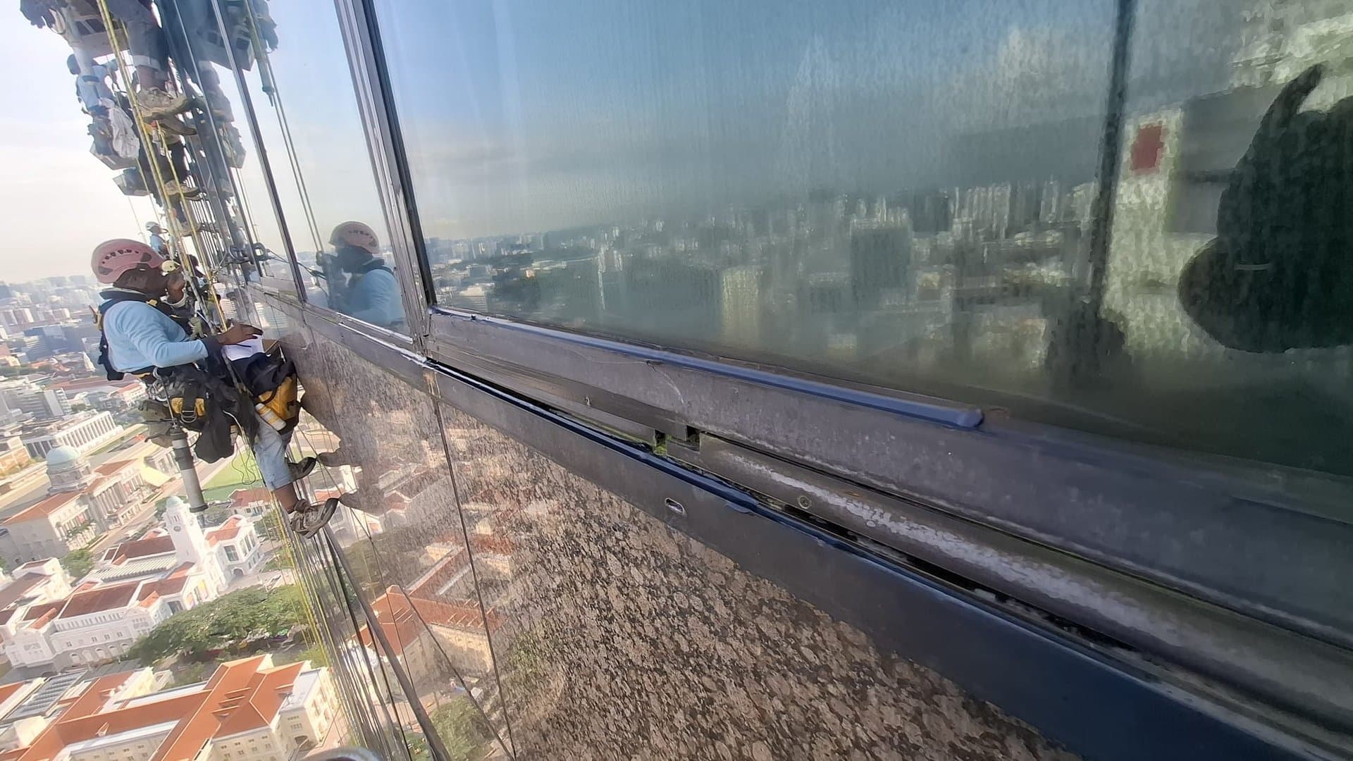 Hydroseal IRATA rope-access technician in full harness and PPE replacing façade sealant on a high-rise office tower in Singapore’s CBD, with the city skyline and river far below